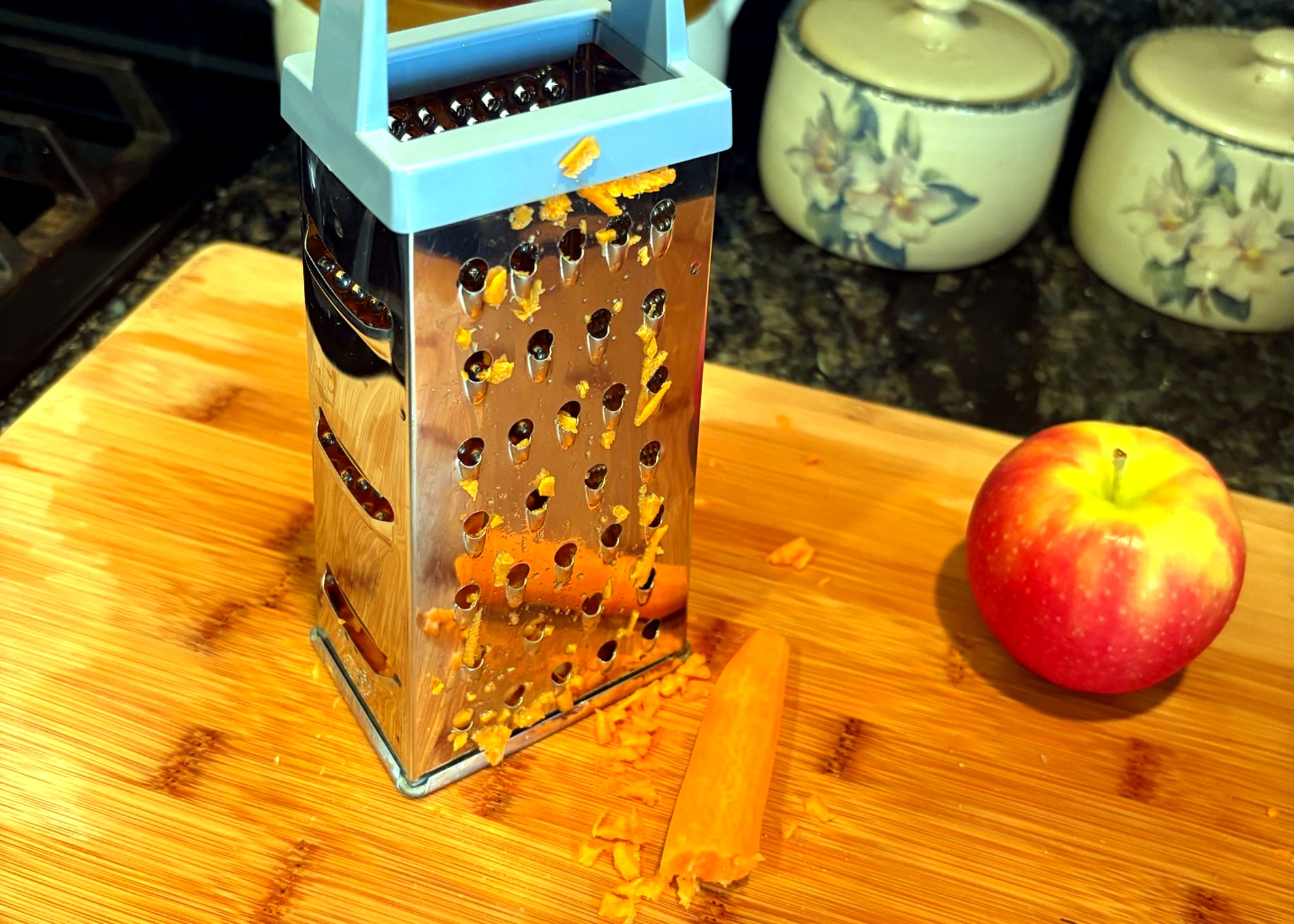Box grater with fresh carrot shreds on a cutting board; apple in background.