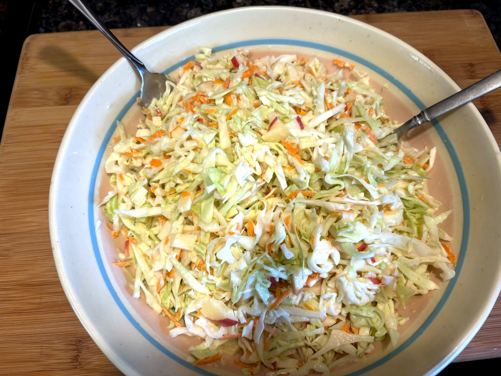 Cabbage, carrot, and apple slaw tossed with creamy cider dressing in a wide bowl; two spoons for mixing.