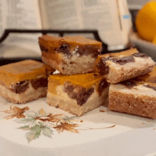 Pumpkin cheesecake chocolate chip bars stacked on a vintage autumn plate, showing distinct layers of shortbread crust, cheesecake, chocolate, and pumpkin custard.